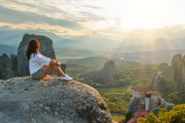 Travel Concept: A dark-haired woman in a white dress sits on a Meteora cliff watching the sunset over the valley with the Meteora monasteries complex.  