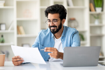 Indian Male Freelancer Sitting At Desk With Laptop And Working With Papers
