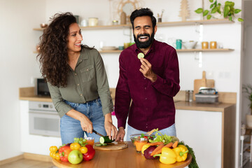 Happy young arab couple cooking healthy meal together in kitchen interior and laughing, man eating slice of cucumber