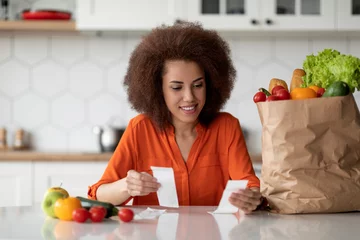 Fotobehang Market Delivery. Happy Black Lady Checking Bills In Kitchen After Grocery Shopping © Prostock-studio