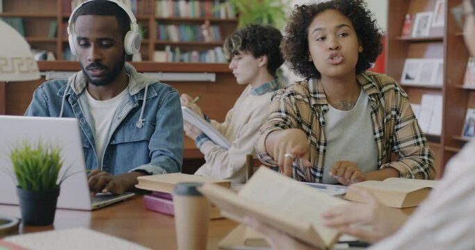 Diverse group of students doing assignments in library reading books and using laptops sitting at desk together. Education and multiculturalism concept.