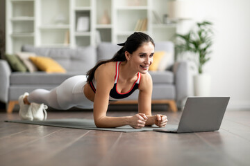 Fototapeta na wymiar Sporty young lady planking at home, looking at laptop screen