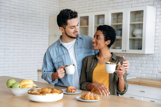 Coffee time in the morning. Happy spouses of different nationalities have tasty weekend breakfast in the kitchen at home with croissants and coffee, spend time together, looks at each other, smile