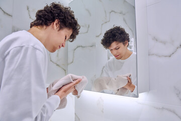 Young guy in bathroom near mirror washing face, wiping with towel