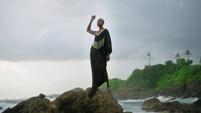 Gay bipoc man in luxury dress, boutique brass jewelry, makeup poses gracefully tropical rocky hill top on ocean beach. Homosexual ethnic elegant person standing on stone like Oshun orisha. Cloudy sky.