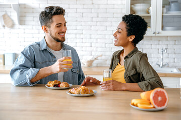 Obraz na płótnie Canvas Happy couple in love, an african american woman and a caucasian man, sit at home in the kitchen, eat croissants for breakfast and drinks juice, enjoy a joint morning together, smile