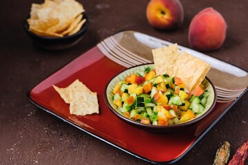 Salsa from fresh cucumber and ripe peach in a ceramic plate on a brown concrete background. Served with wheat chips. Mexican cuisine.