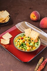 Salsa from fresh cucumber and ripe peach in a ceramic plate on a brown concrete background. Served with wheat chips. Mexican cuisine.