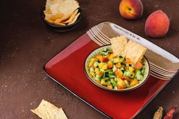 Salsa from fresh cucumber and ripe peach in a ceramic plate on a brown concrete background. Served with wheat chips. Mexican cuisine.