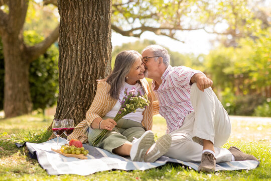 Smiling old european husband gives bouquet of flowers to wife, kiss, enjoy picnic together in park