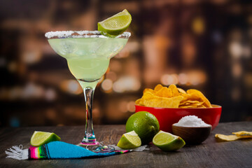 Margarita cocktail with sliced and whole limes, nachos and salt on wooden bar table. Mexican party background
