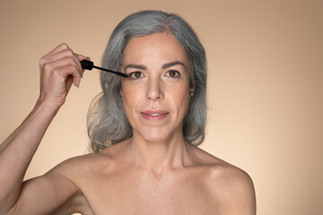 Portrait of senior woman applying mascara on her lashes, putting on decorative cosmetics, beige background, free space