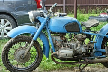one old rusty iron classical  heavy big comfortable blue   dirty made in the ussr retro  motorcycle stands on green grass  near the car  on the street during the day