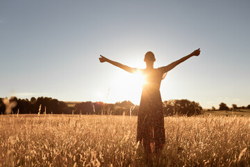 Young female adult in field, arms raised, embracing sunlight and happiness.
