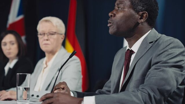 Medium side shot of African American politician in formal suit and tie sitting at table at international symposium and speaking persuasively, while Caucasian female colleagues are looking on