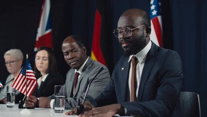 Medium shot of African American prime minister in glasses giving speech on current agenda at international symposium, and diverse colleagues listening, with UK, USA and German flags in background
