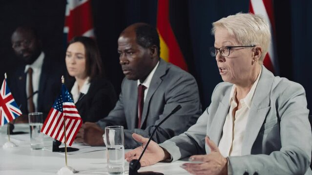 Medium shot of middle-aged blonde Caucasian female politician presenting serious argument at international summit, diverse politicians listening, and flags of USA, UK and Germany in background