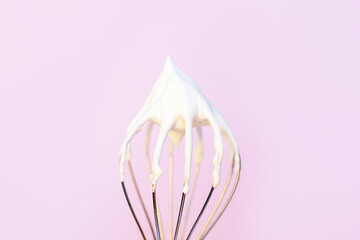 Whisk with whipped cream on light pink background close-up.