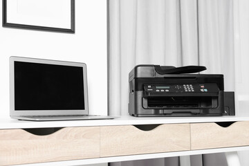 Modern printer with paper near laptop on white desk at home