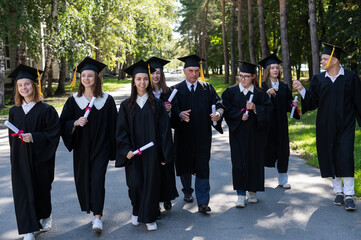 A group of graduates in robes with diplomas in their hands walk outdoors. Elderly student.