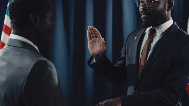 Medium tilting shot of bearded middle-aged bearded black male politician in suit, tie and glasses giving oath on holy Bible or Quran, with raised hand, American flag and dark blue drapes in background