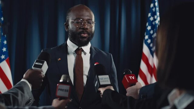 Medium shot of African American male politician or presidential candidate, in suit, tie and glasses, with vote badge, speaking in front of journalists with microphones during election campaign