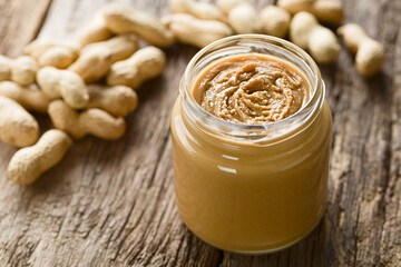 Creamy smooth peanut butter in jar with peanuts in shell in the back, photographed on wood (Selective Focus, Focus in the middle of the peanut butter)
