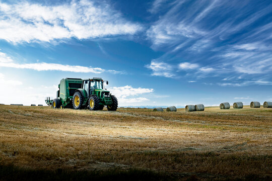 A tractor pulling a baler across a harvested field with round straw bales at background under a deep blue sky in Rocky View County Alberta Canada.