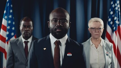 Chest-up portrait shot of charismatic African American presidential candidate and multiethnic male and female advisers posing in front of USA flag during election, raising head and looking at camera - Powered by Adobe
