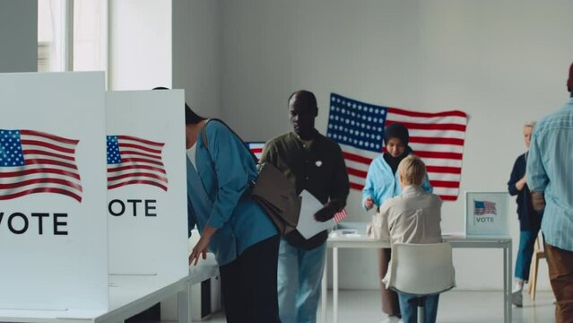 Full shot of diverse male and female American citizens walking up to election official at polling station, taking ballot paper, filling it in small cubicle, stuffing into ballot box and leaving