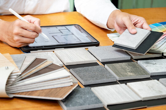 Architect hand choosing and picking stone and wood material samples while drawing on a digital tablet on the table in studio. Designer working for interior architecture and furniture design project.