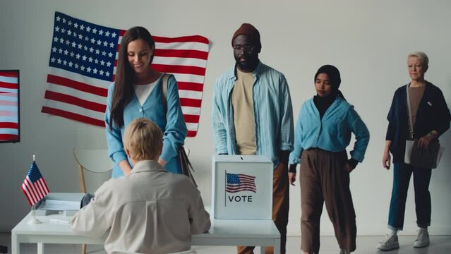 Full shot of group of casually dressed diverse Americans standing in queue at election precinct, walking up to official, greeting, taking ballot and walking away towards booths