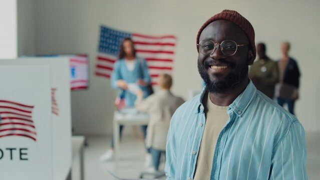 Medium close-up portrait shot of young African American man in beanie, glasses, denim shirt smiling happily, after voting at polling station at federal elections, diverse people queuing in background
