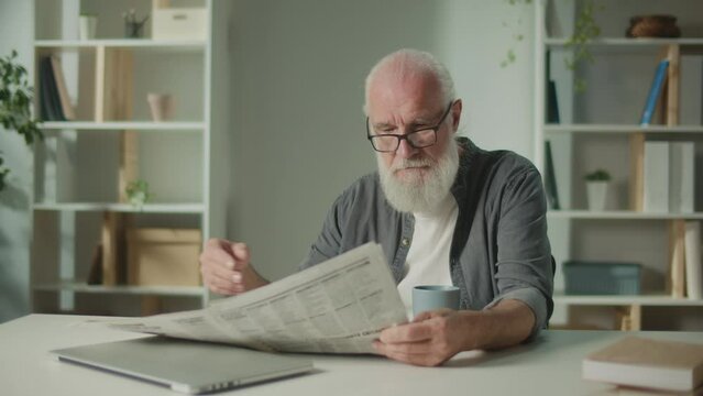 A Serious, Smart Old Man Sits at a Table and Reads a Newspaper.An Elderly Man with a Newspaper, Drinking Coffee Alone at Home, Sees the News and Current Events in the Newspaper.Daily Newspaper Concept