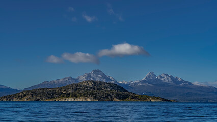 Naklejka premium A picturesque mountain range of the Andes against a background of blue sky and clouds. In the foreground is the blue water of Ensenada Bay. Argentina. Tierra del Fuego National Park. Ushuaia.