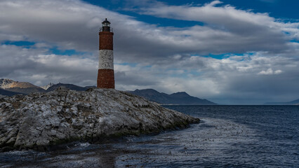 An ancient lighthouse stands on a rocky island in the Beagle Channel. A red-and-white striped tower against a  blue sky and clouds. Argentina. The world's southernmost lighthouse Les Eclaireurs