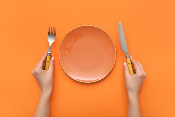 Female hands with cutlery and empty plate on orange background