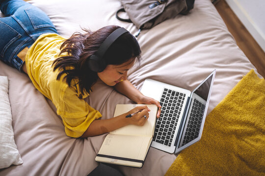 Top view of teen Asian college girl wearing headphones lying on bed planning week on planner. Studying using laptop. - Powered by Adobe