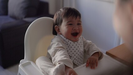 Back view of mom feeding her baby. Toddler is sitting on a high chair and eating with spoon