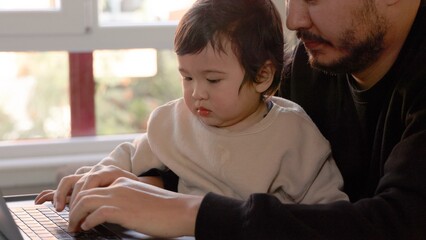A young man trying to work on laptop holding his son at home. Remote work