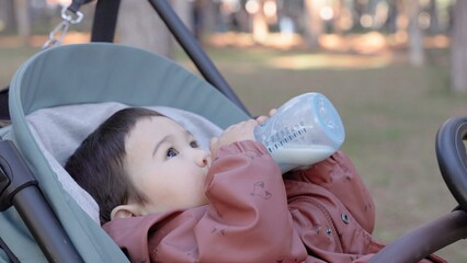 Asian baby drinks formula from a bottle while lying on the stroller in the park