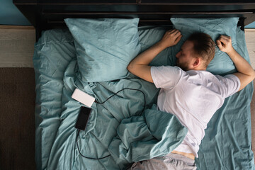 Top view of millennial man sleeping on the bed and turning off smartphone alarm clock in the...