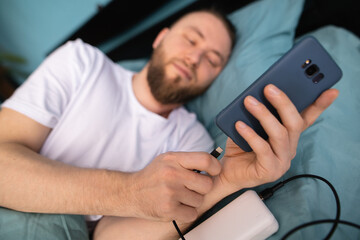 Serious millennial man using mobile phone in bed connect it to a charger while lying on his side