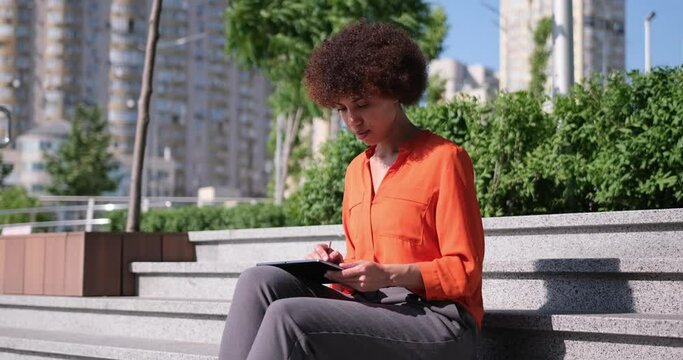 Happy young African American woman using digital tablet pad sufring internet inspired with new ideas sitting outdoors