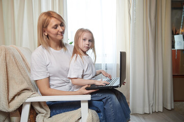 Beautiful young woman and her little cute daughter are using laptop at home. Enjoying spending time together with Internet and modern technologie