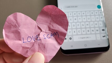 Love scam concept. A broken heart victim holding a crumpled heart with the words Love Scam written. Off focus background with a mobile phone message to the scammer  - obrazy, fototapety, plakaty