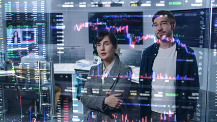 Team of stock traders look at computer generated virtual 3D exchange market charts on glass wall, analyze business strategy in broker agency office. Computers and big digital screens on background.