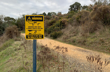 black and yellow Warning: Dangerous Mine Shafts and Tunnels Occur in this Area - Keep Clear At All Times metal sign on post near walking track - obrazy, fototapety, plakaty