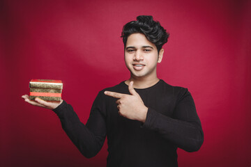 Gift Concept Young man pointing at Valentine's gift copy space isolated on red studio background. A young man, radiating enthusiasm and joy, stands in a vibrant red studio. With an eager expression.