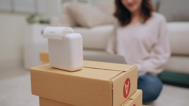 Young asian woman selling online receiving order via laptop and using label printer to printing customer address attach to the parcel box. Business Online and delivery shop concept